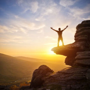 Un homme levant les bras au ciel sur une montagne face au soleil